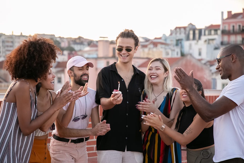 Group of positive, extroverted, young multiracial friends clapping and smiling while celebrating birthday of happy guy making wish with cupcake in hand on building rooftop
